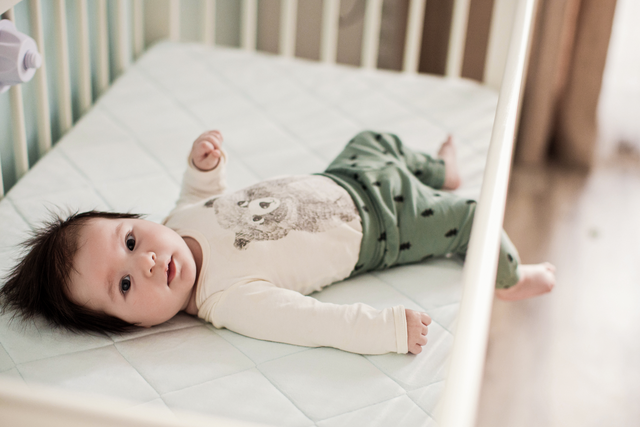 a 6 month old baby with fuzzy dark hair lays in the crib with one leg hanging out. 