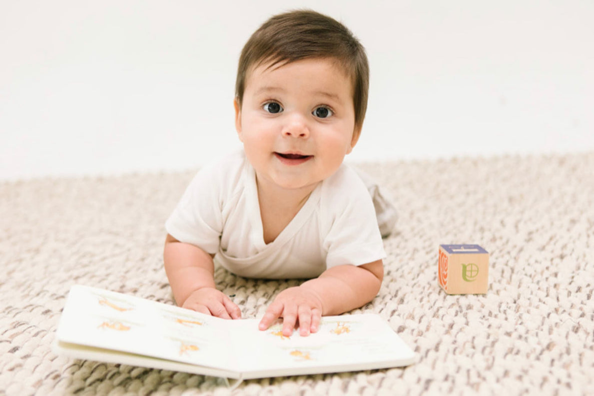 an 8-10 month old baby lays on their tummy on a soft cream colored rug reading a book with a block next to them.