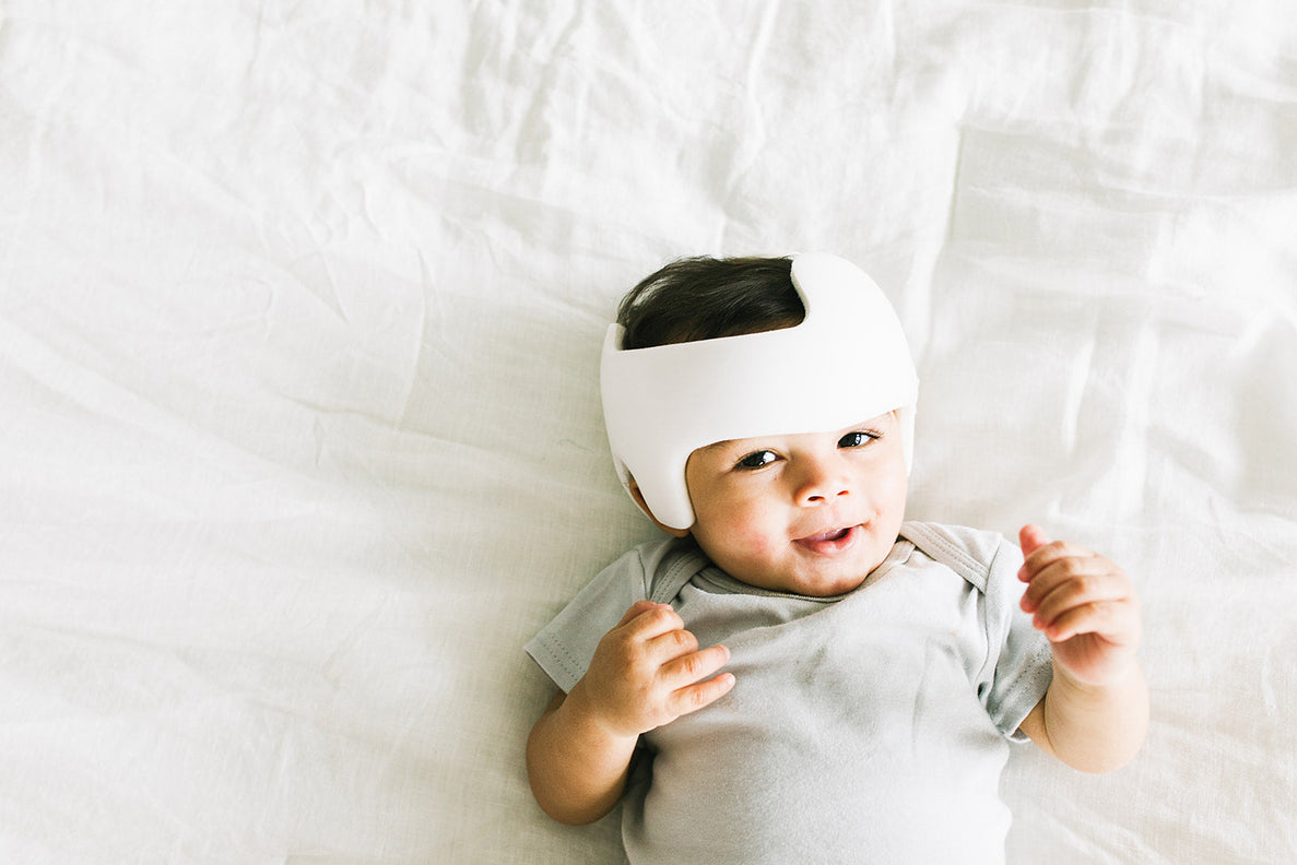a  9 month old boy wearing a DOC band helmet lays on a white sheet smiling at the camera