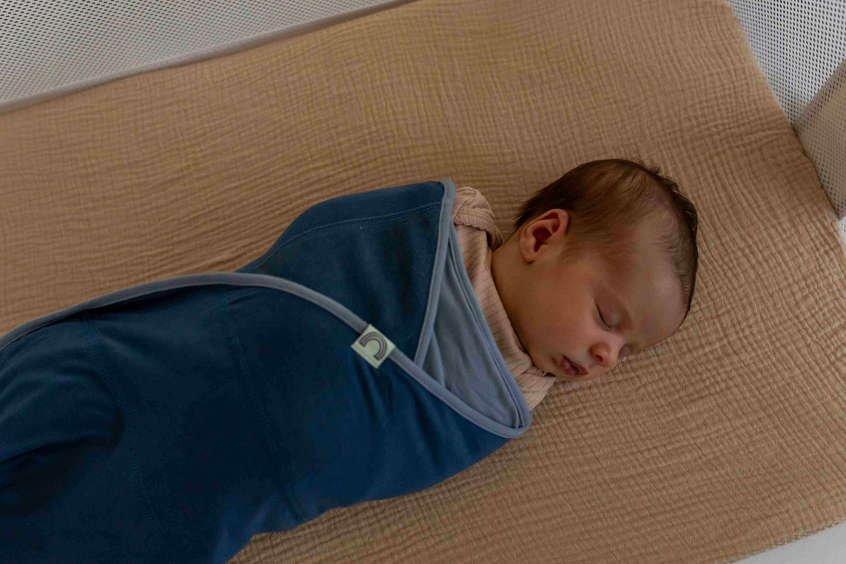 A newborn in a blue swaddle sleeps on a flat peach colored surface