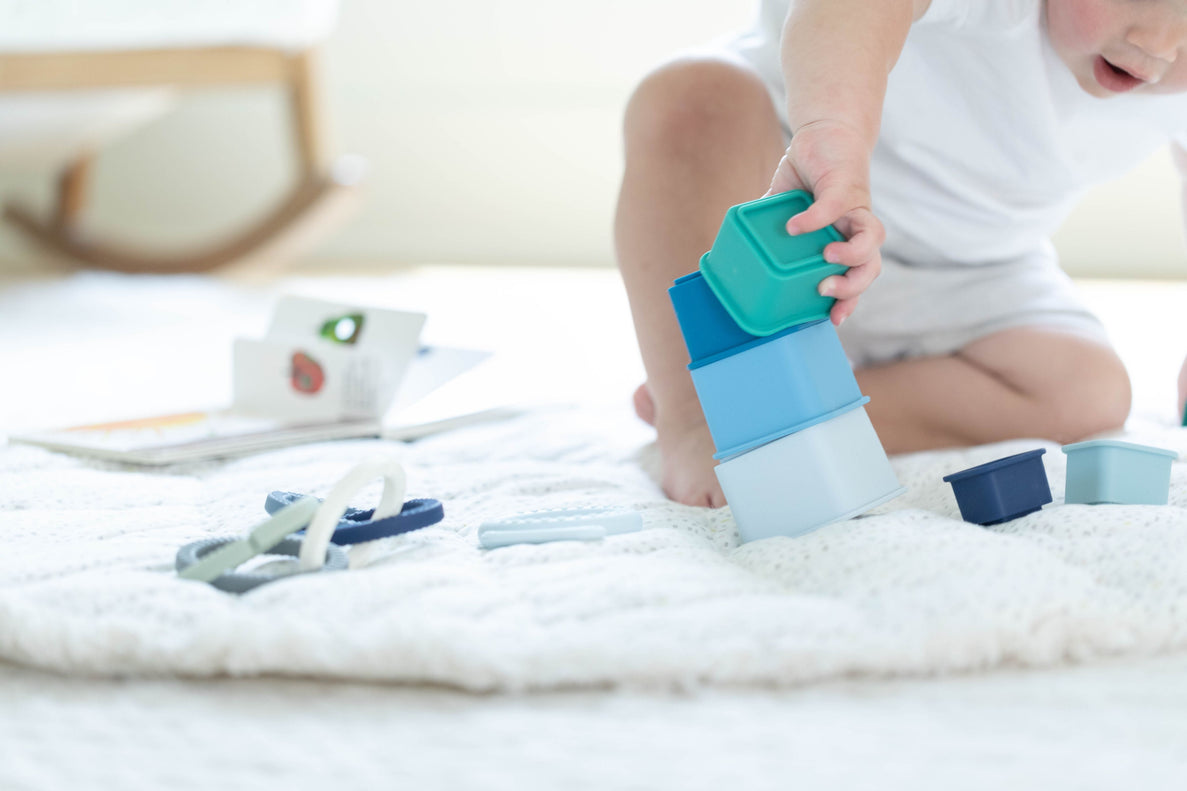 a baby plays with a blue stacking toy while sitting on a white fluffy rug with toys and books around them.
