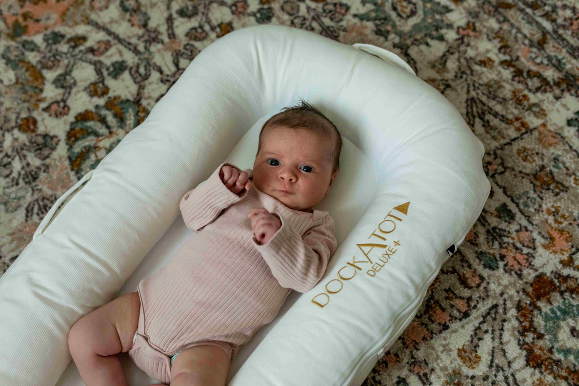a newborn baby in a pink onesie lays in a DockATot that is on top of an oriental patterned rug