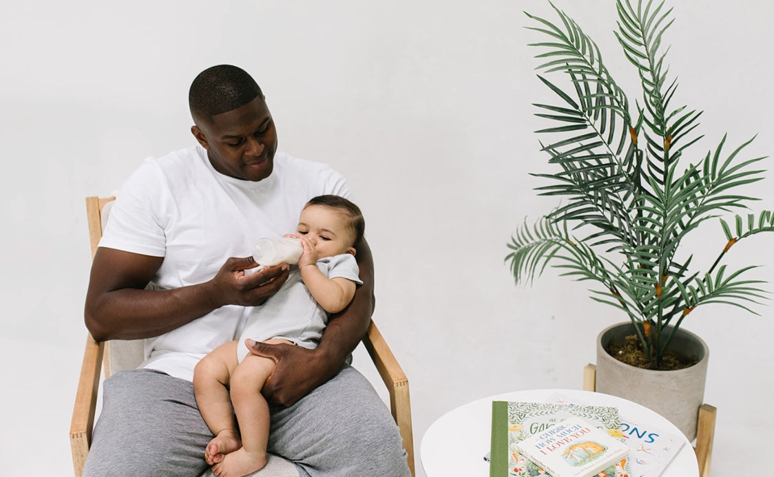 A dad sits in a chair next to a table with children's books on it and a potted plant. He is feeding a bottle to a baby wearing a onesie
