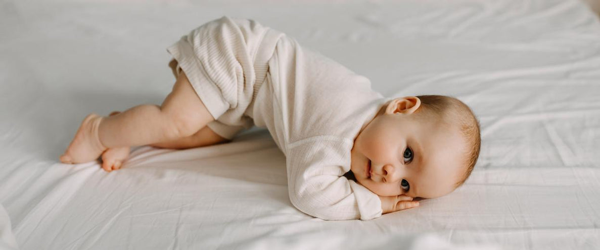 A baby in a light-colored romper is lying on its belly on a white sheet, with its head resting on its hands and knees bent, looking directly at the camera