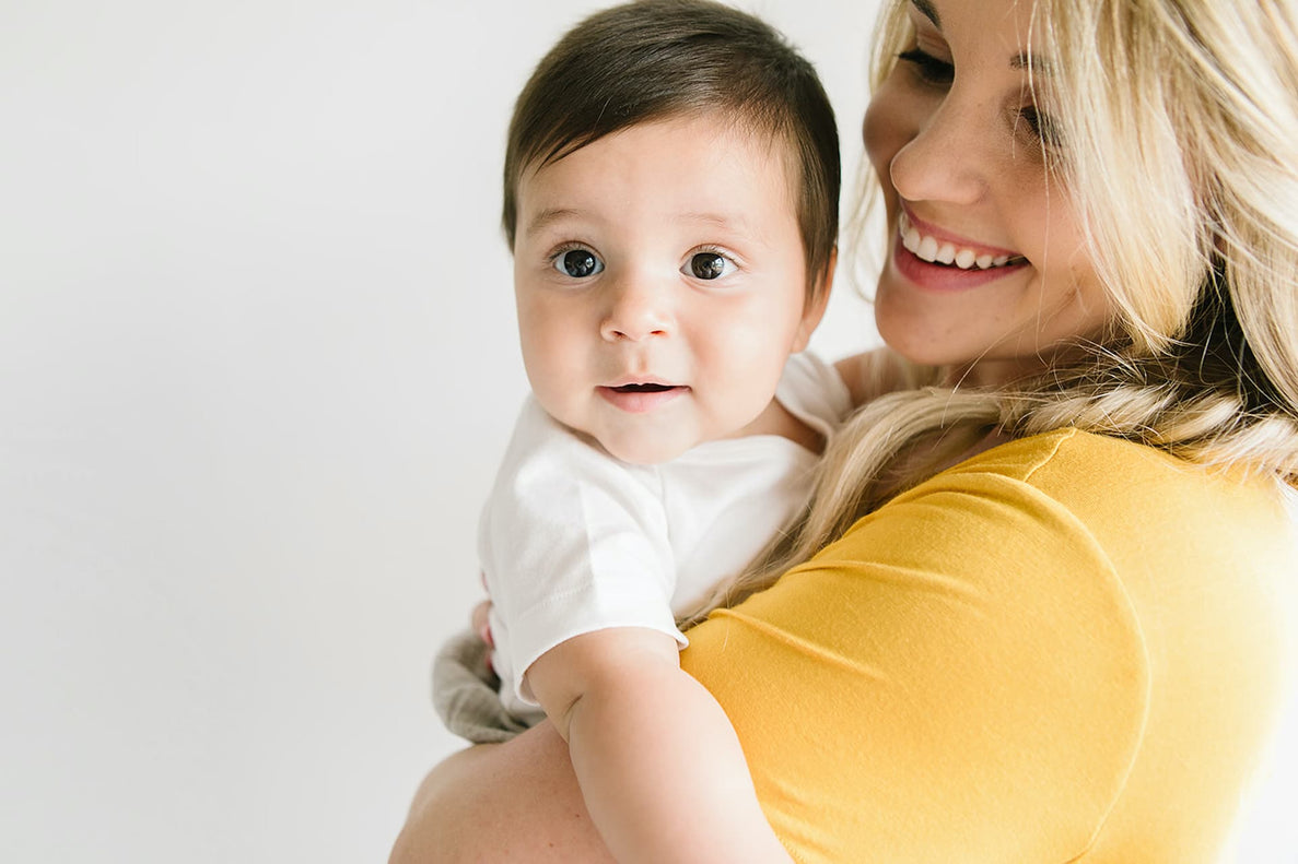  A babysitter with long blonde hair smiles while holding her baby. The baby, who has big dark eyes and is wearing a white onesie, looks directly at the camera with a surprised or curious expression