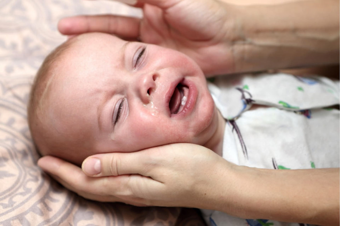 a closeup of a sick baby crying while adult hands try to comfort them. The baby's nose is runny