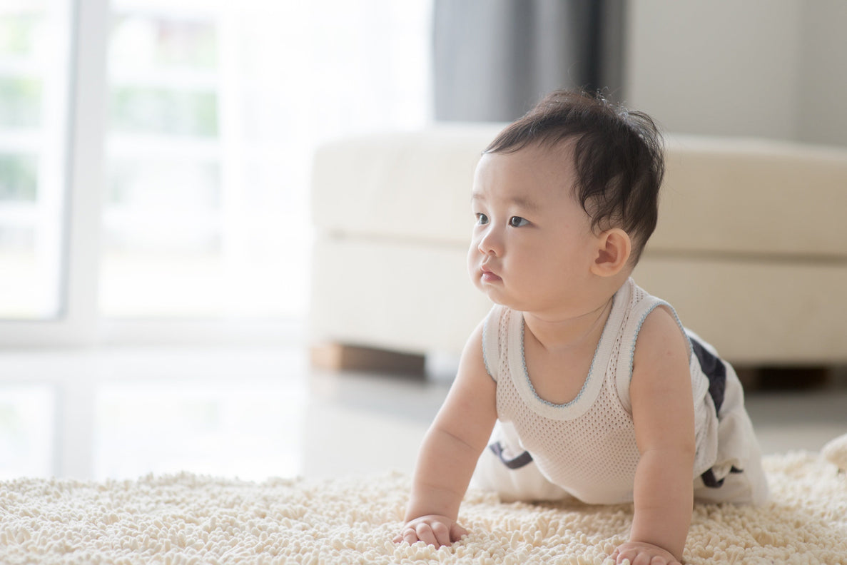 a baby crawls on a white fuzzy rug. He is wearing a white tank top and patterned bottoms. His hair is black. 