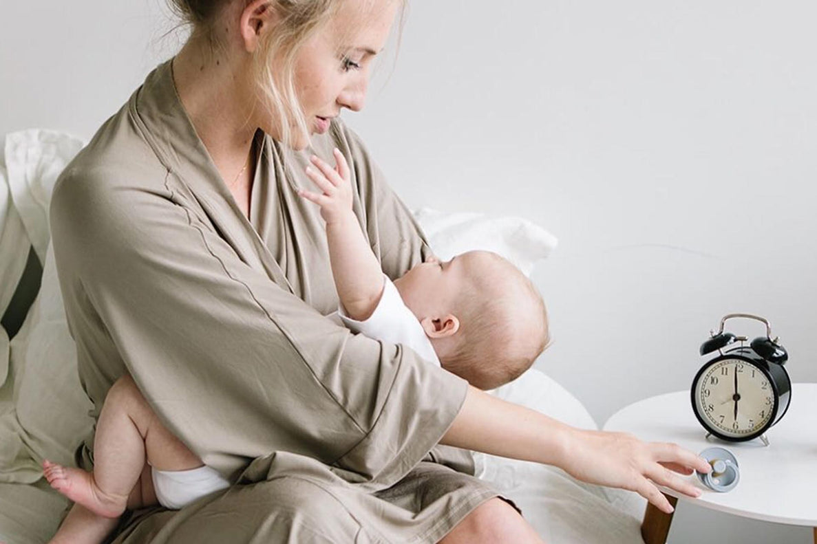 a mother comforts a discontent baby while sitting next to a clock that reads 6:00