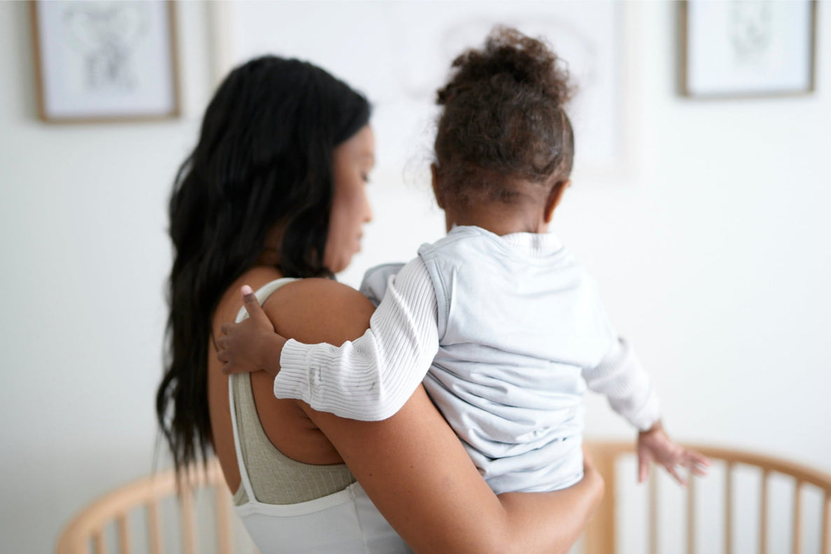 a mom holds an older baby as they stand next to a crib. Baby is reaching for the crib