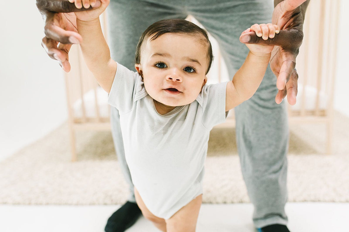 A baby is learning how to walk by holding an adult's hands. A crib is in the background