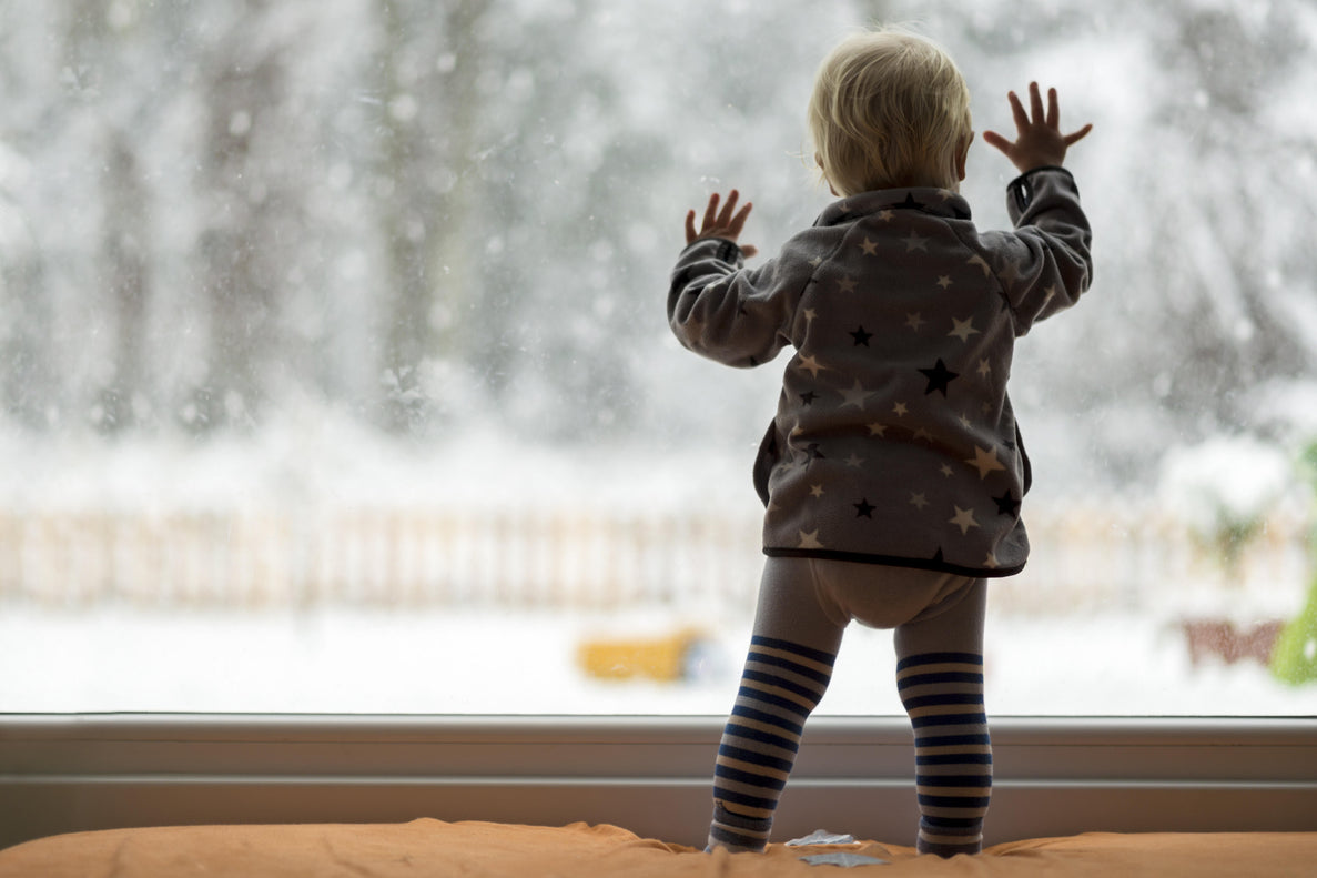 a young toddler baby dressed in cozy clothing has both hands on the window as they look out at the snow coming down