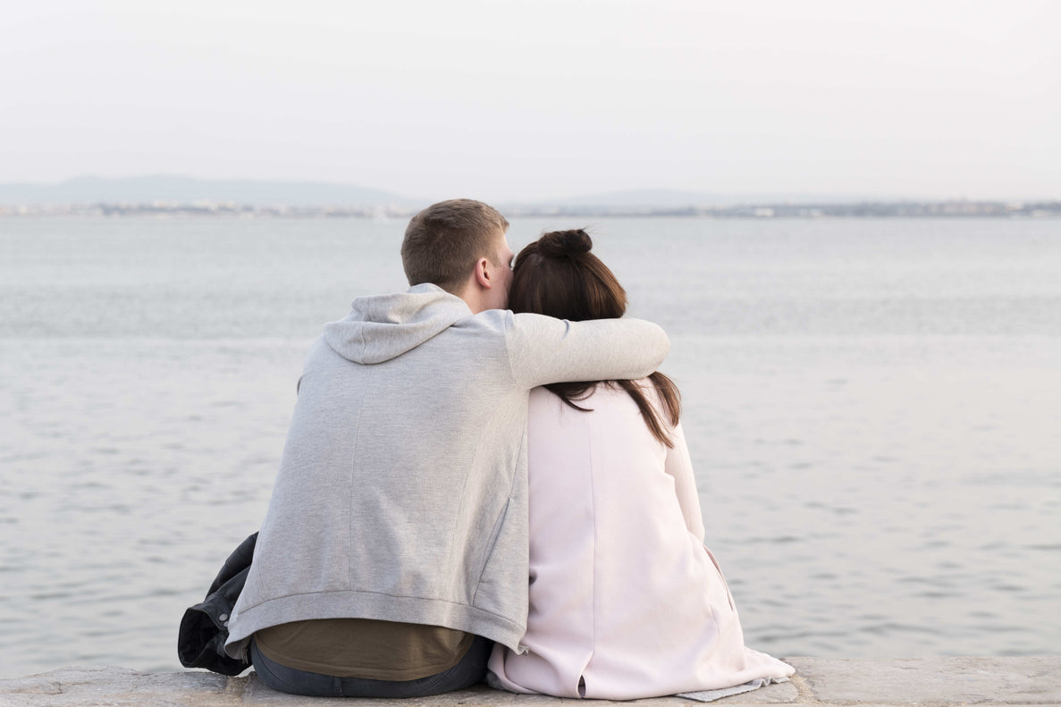 A couple is sitting by a large body of water (like a sea or lake), seen from behind, with the man's arm around the woman's shoulder.