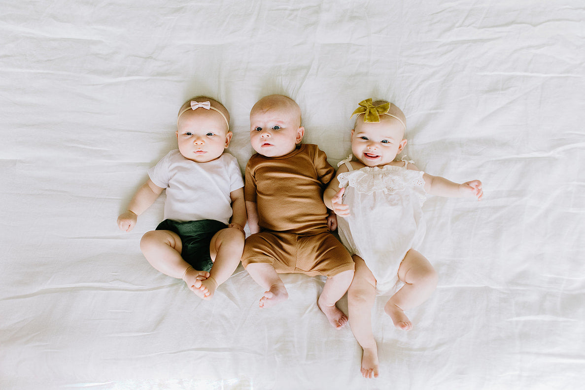 three 3 month old babies lay next to each other on a bed. One is smiling with outstretched arms and 2 are looking more inquisitively 