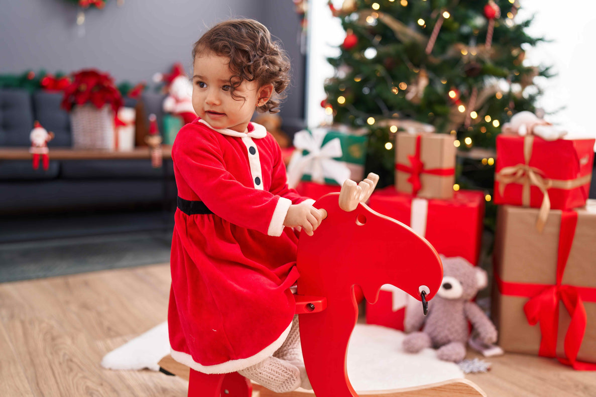 a young girl dressed in a santa dress rocks on a red wooden moose in front of a christmas tree with gifts