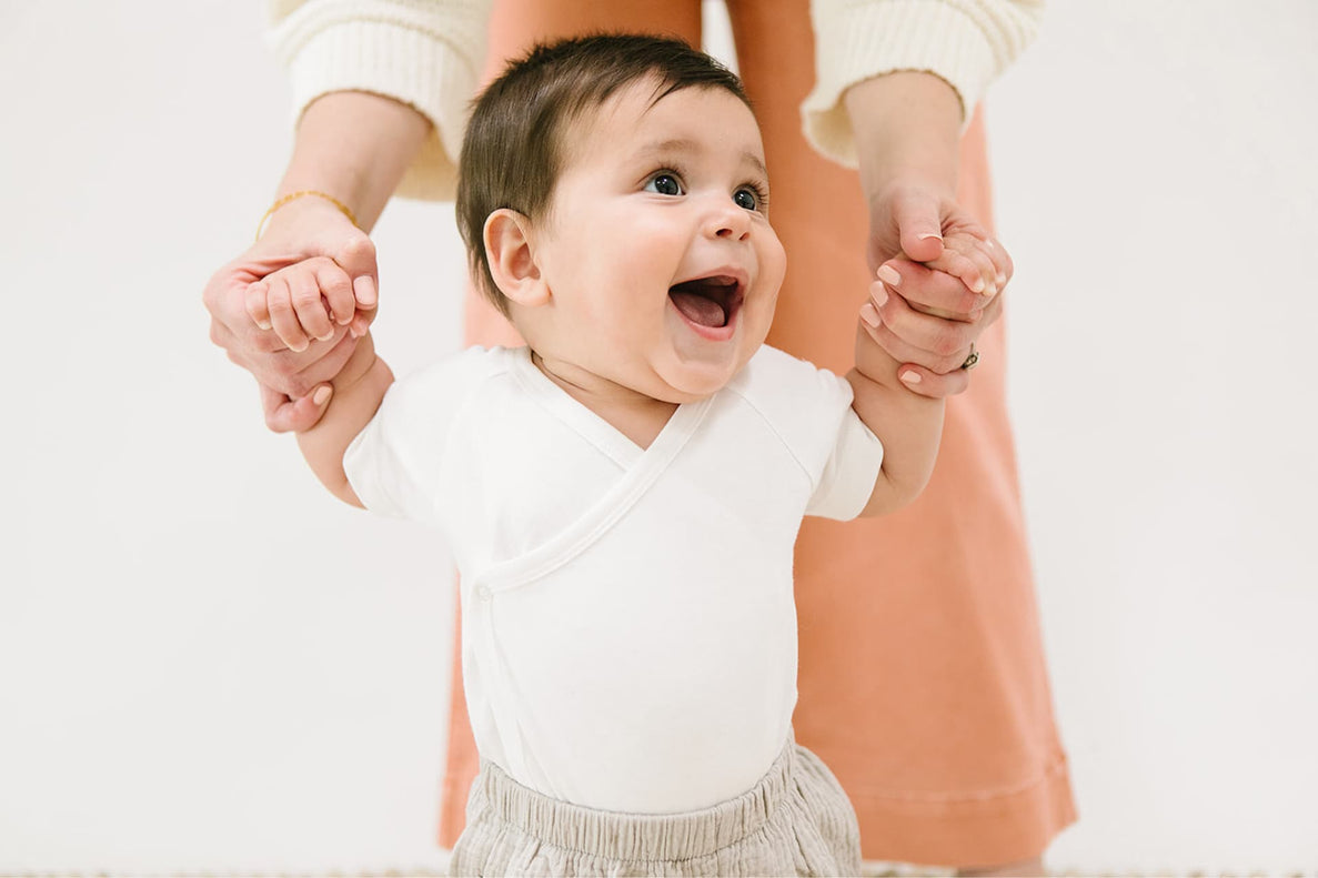 A baby learns to walk by holding on to an adults hands. The baby has an excited look on his face