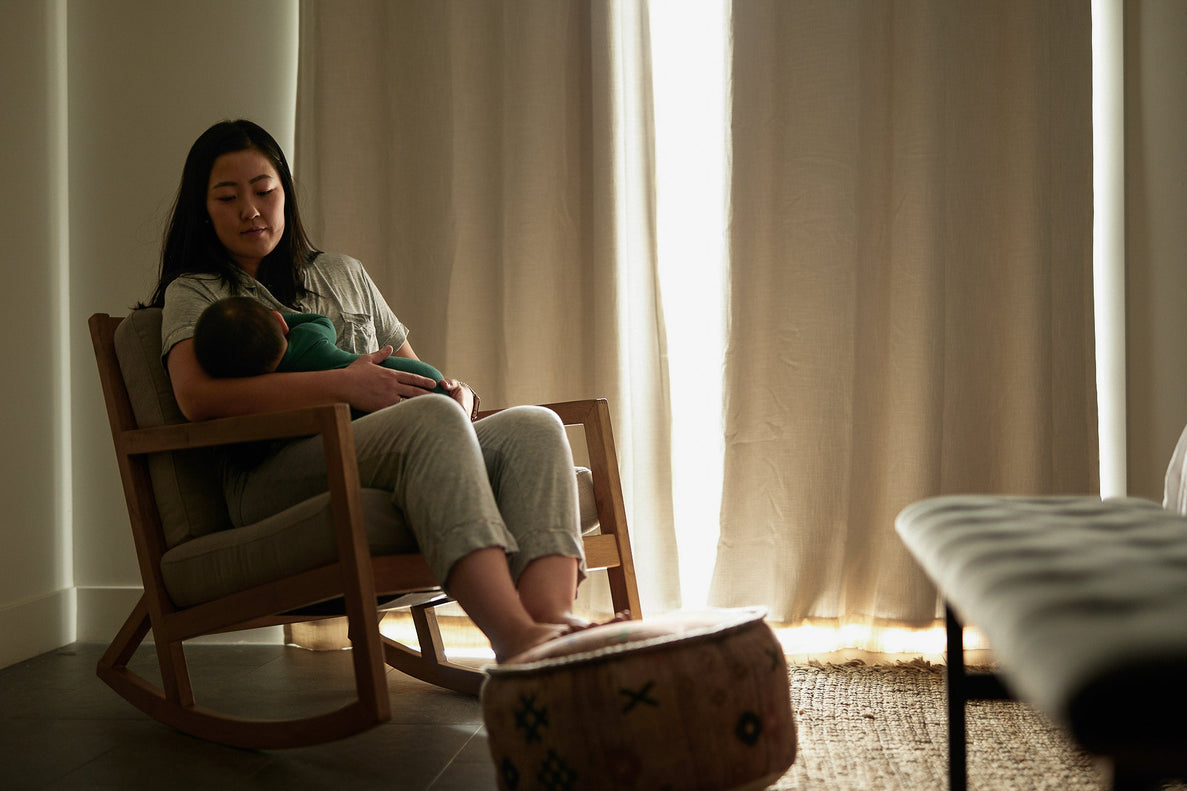 A mom sits in a rocking chair in a darkened room with light peeking through the curtains. She is holding a sleeping baby in her arms