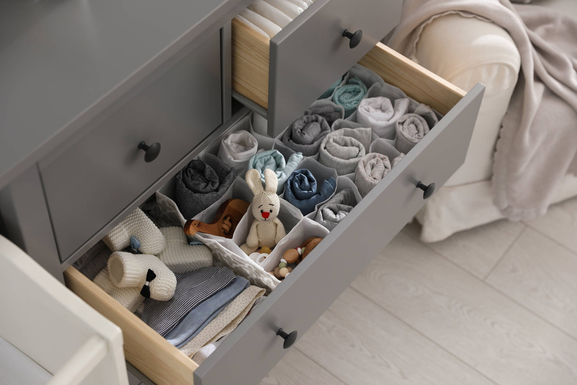 a closeup of chest of drawers in a nursery. The bottom drawer is open and contains neatly organized baby clothing and other items