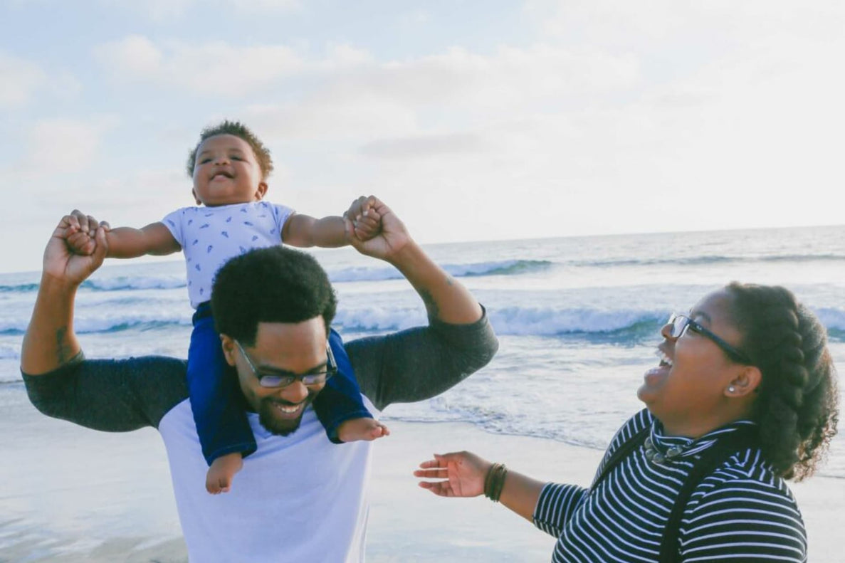 a mom, dad, and baby walk along the shore of a beach. They all smile as baby rides on dad's shoulders
