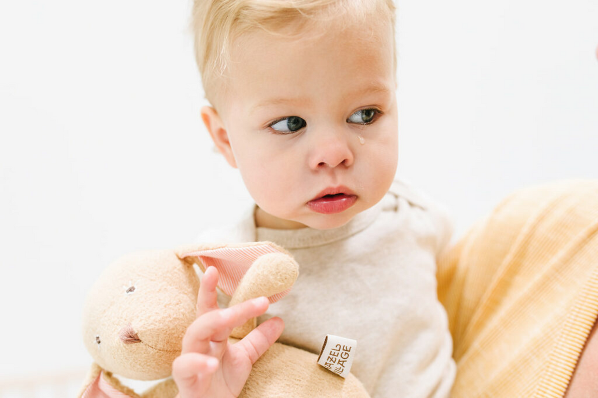A close-up of a fair-haired toddler in a neutral-colored shirt, with a tear running down the cheek, clutching a soft, brown bunny toy