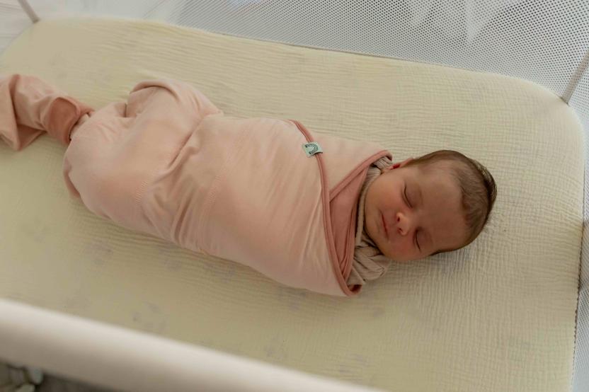  A sleeping newborn baby, wrapped tightly in a pink swaddle with a small teal snap or clasp, lies on its back on a lightly patterned, pale yellow crib mattress inside a white crib with mesh sides.