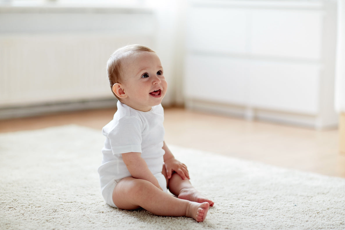 a baby dressed in white sits on a white rug smiling