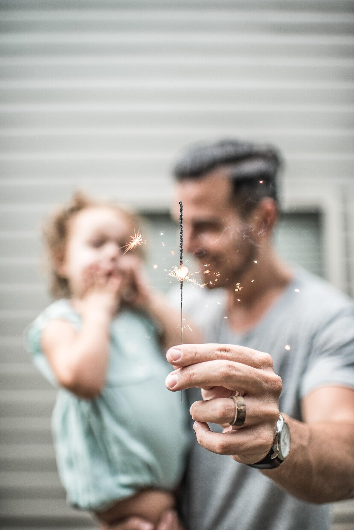 a dad holds his older baby girl in one arm and holds a sparkler out at arms length with the other hand