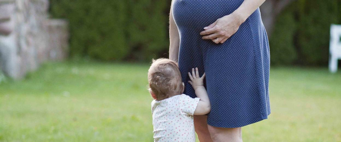 A pregnant mom stands in a green backyard with a toddler clinging on to her legs