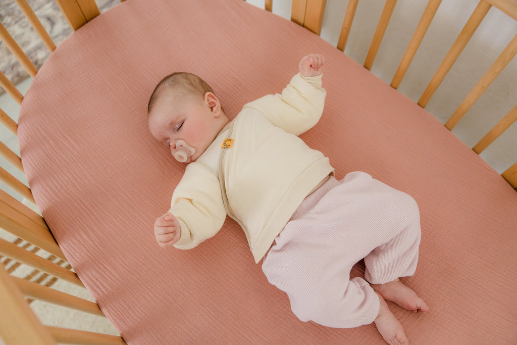 baby wearing coconut cream Jacky swaddle transition jacket layered over pink pajama pants laying on back in a crib