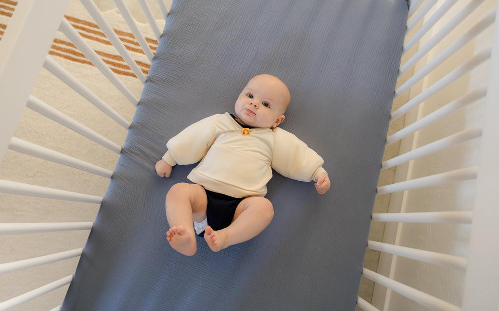 baby wearing coconut cream Jacky swaddle transition jacket layered over black onesie laying on back in a crib