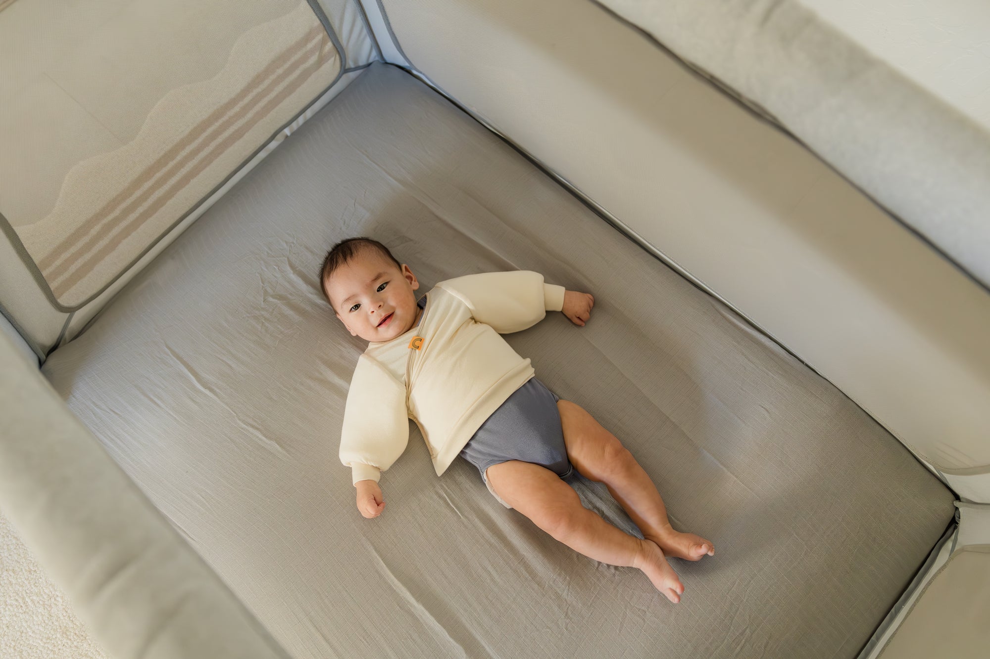 baby wearing coconut cream Jacky swaddle transition jacket layered over gray onesie laying on back in a portable crib